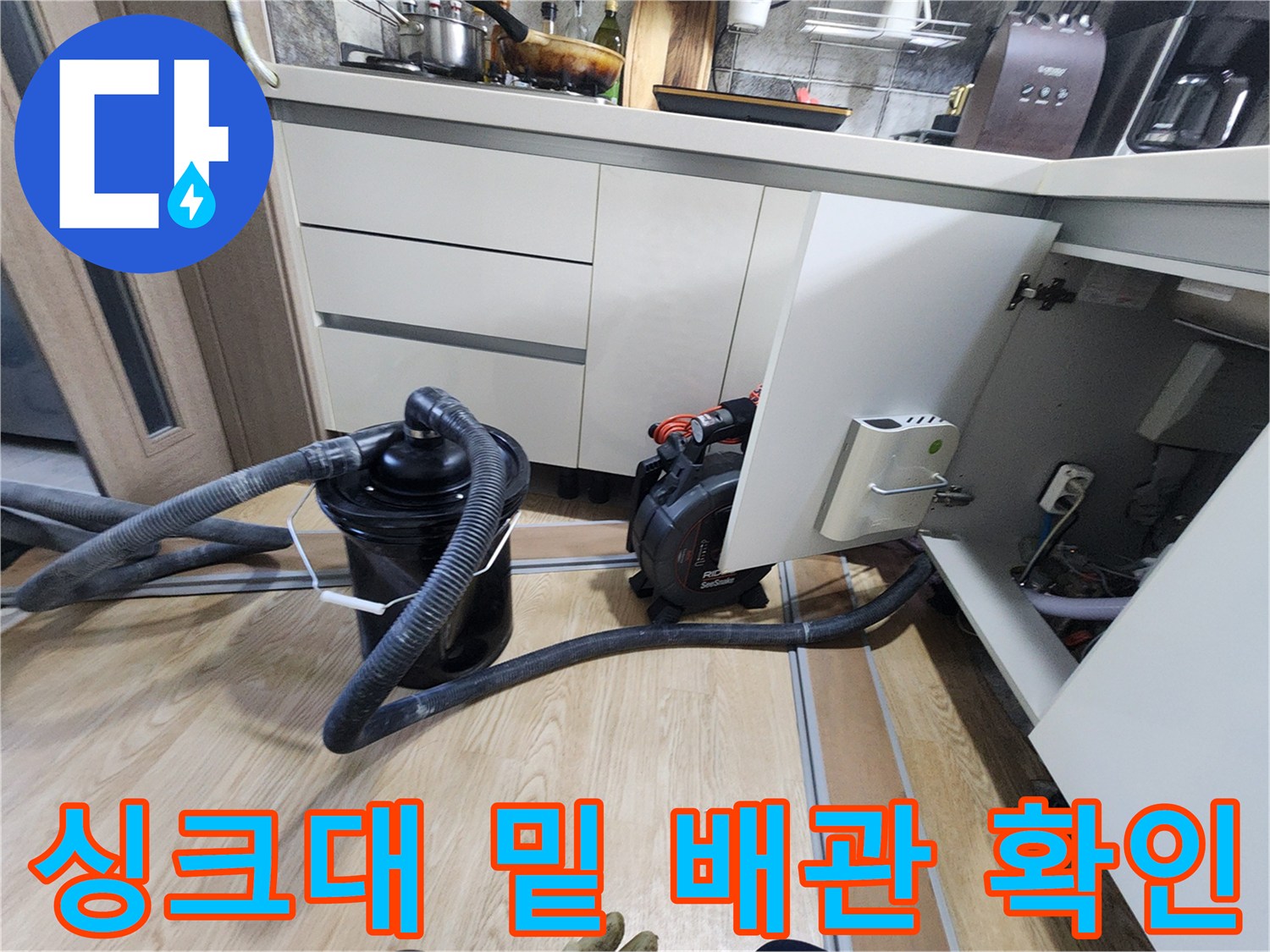 싱크대 하부장 아래 배수 호스와 점검 장비가 놓여 있는 장면입니다.
