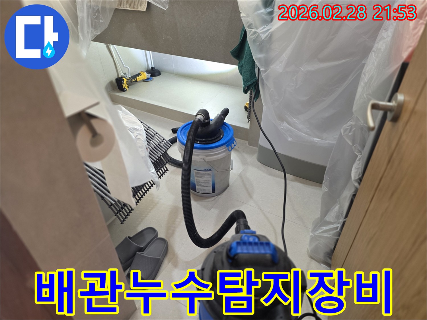 이 이미지는 배관 누수를 탐지하기 위해-습식 청소기와 연결된 특수 버킷 장비를-욕실 내부에 설치하고 작업하는 현장을 보여줍니다.