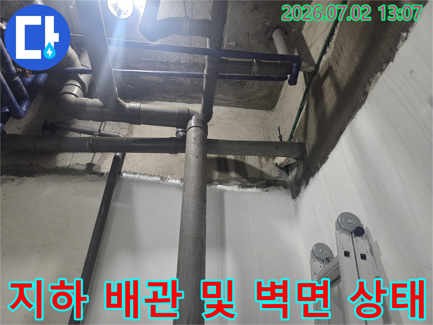 천장에서 수직으로 내려오는 굵은 회색 배관과 습기로 오염된 지하 벽면