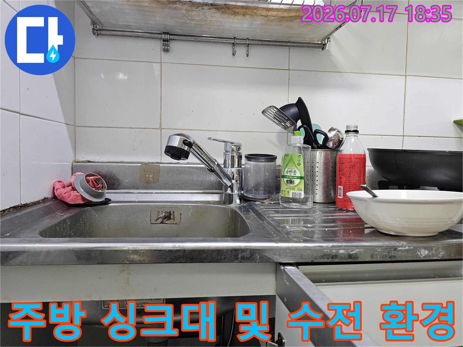 흰색 타일 벽면 앞의 스테인리스 싱크대-수전과 세정제가 놓여있음