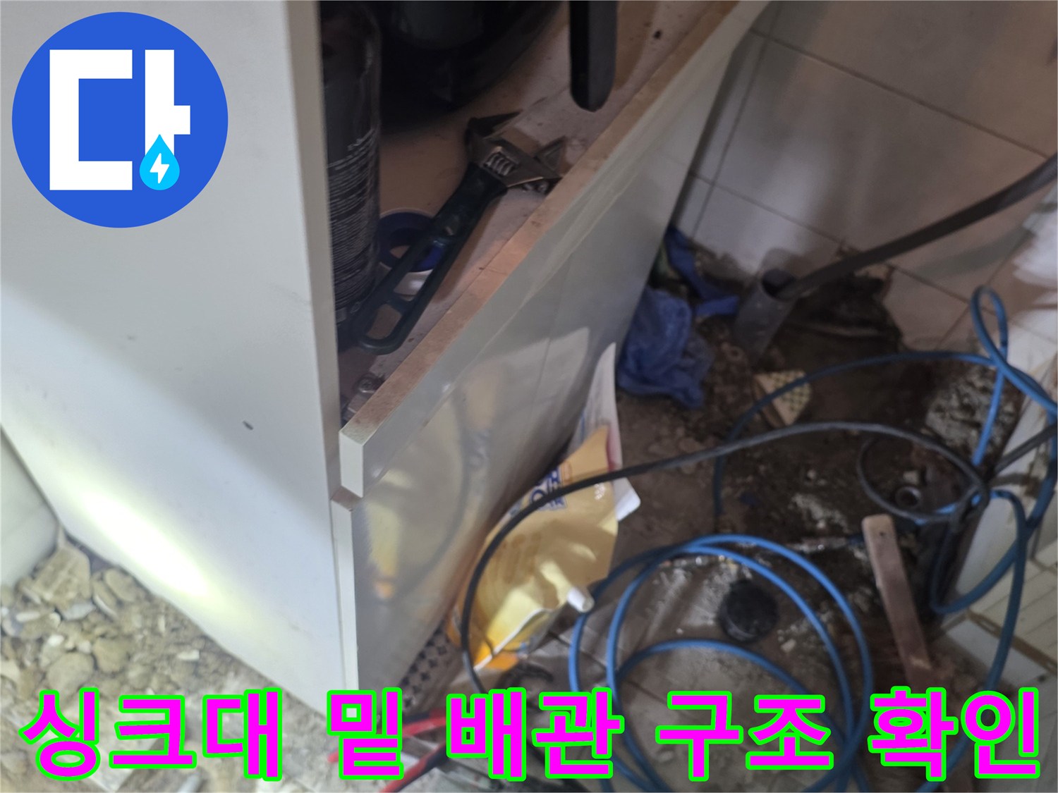 주방 싱크대 아래 복잡하게 얽힌 배관과 작업 도구들의 모습입니다.