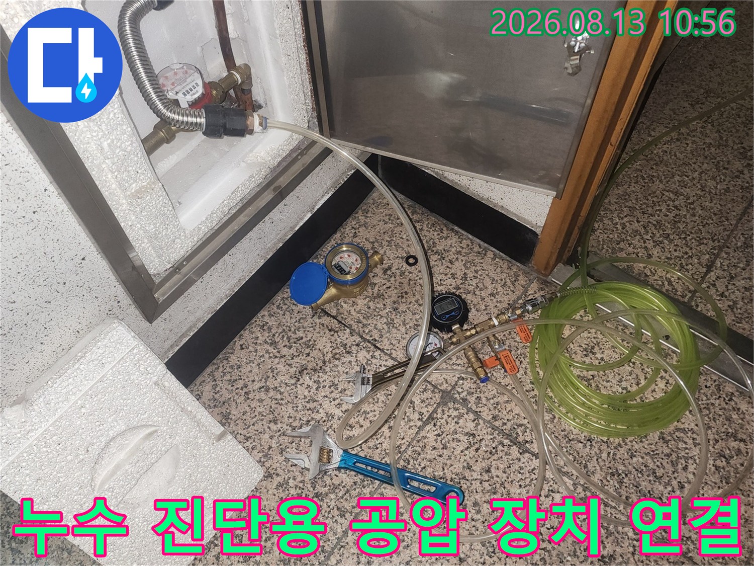 바닥 점검구 내부 배관에 투명한 호스와 압력계가 설치된 모습-작업 도구가 나열됨