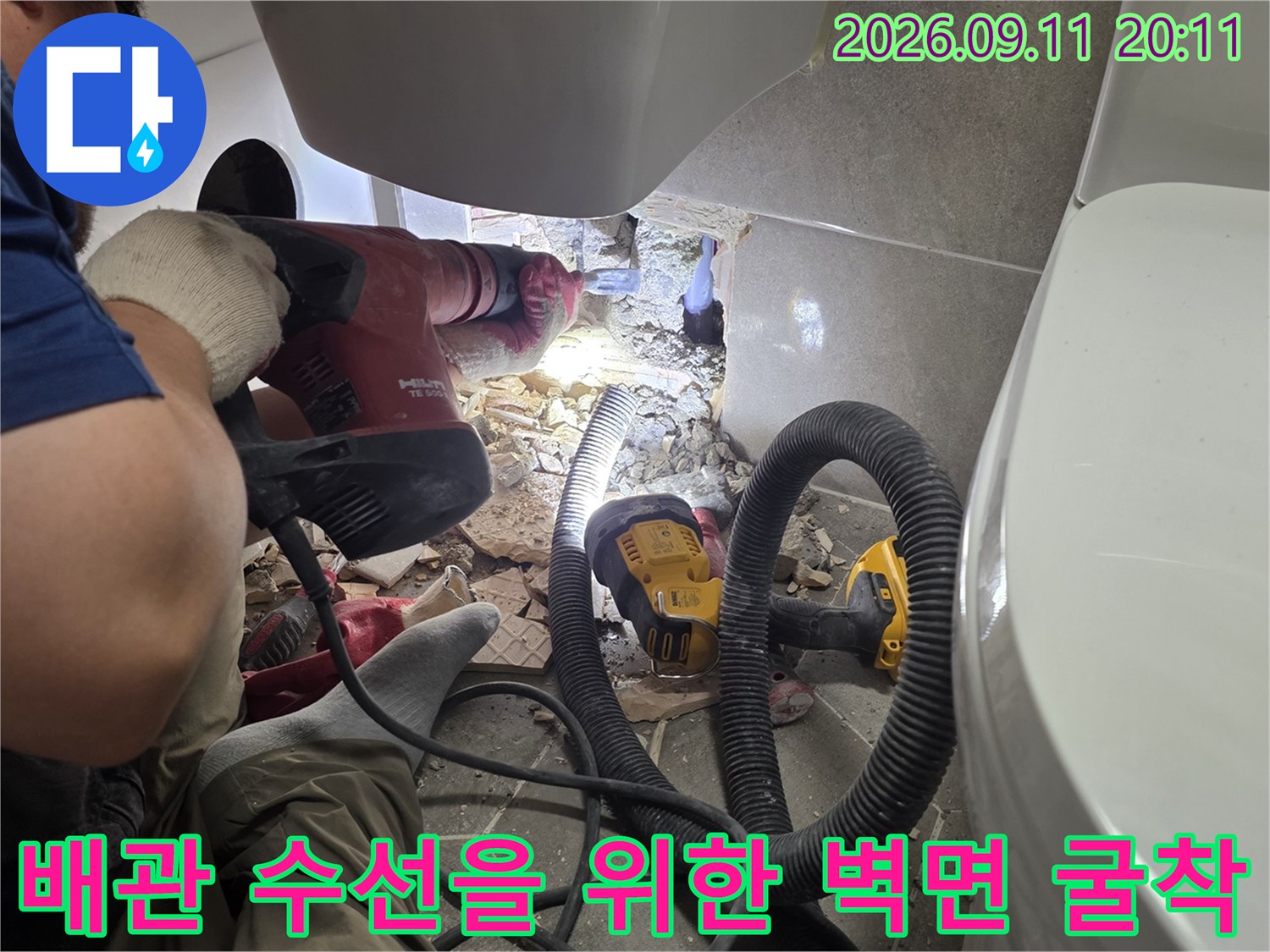 전동 드릴과 흡입 장비를 사용하여 세면대 밑 배관 부위를 파내는 모습입니다.